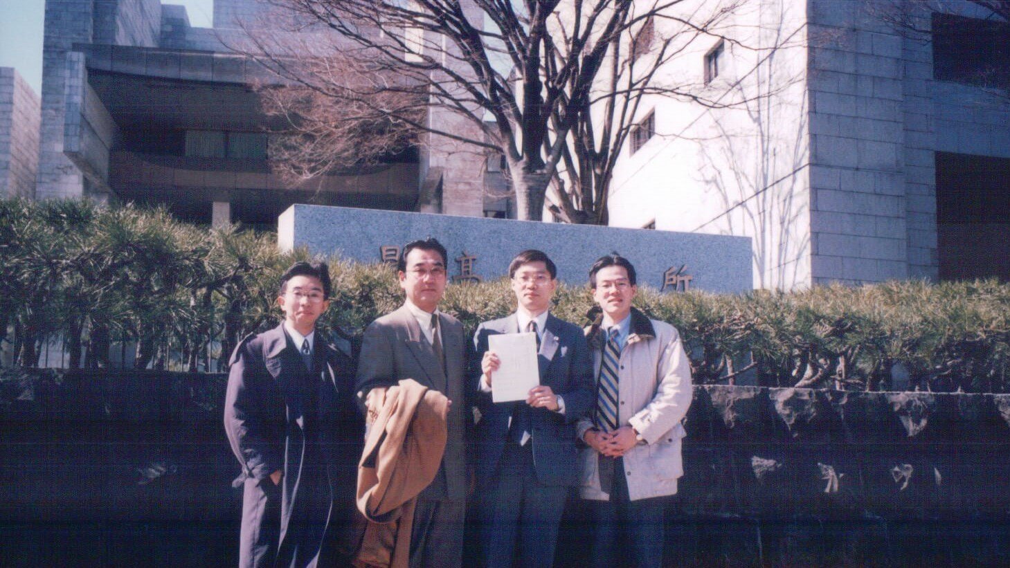 four men standing in front of a wall with buildings in the background holding a document